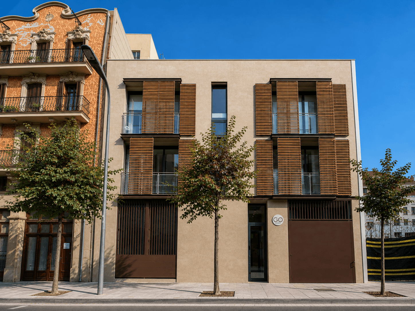 Edificio de tres plantas en una calle arbolada, revestido en tono arena, con contraventanas de lamas de madera y balcones de barandilla de vidrio.