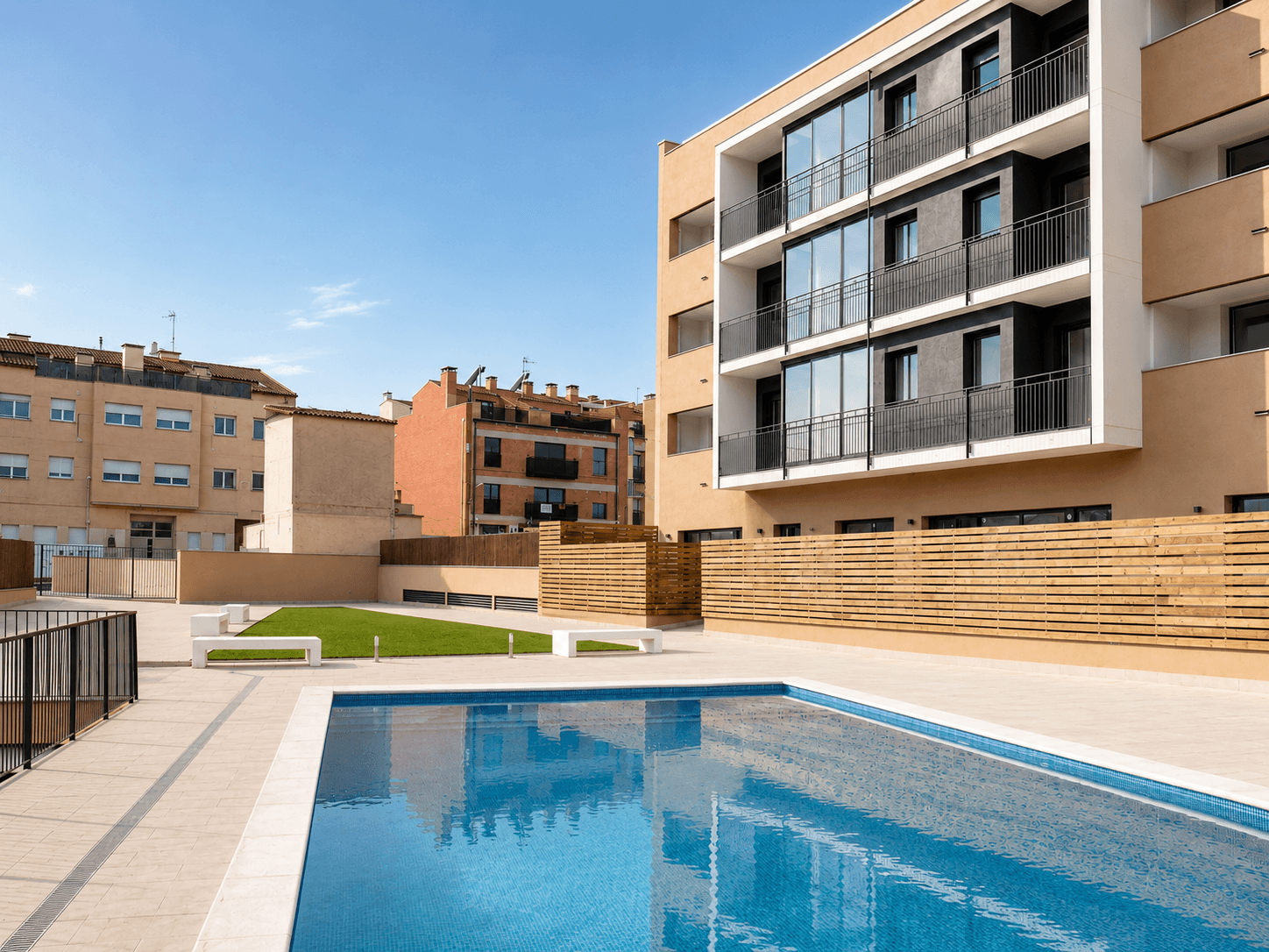 Piscina comunitaria con césped y vallado de listones de madera junto a un bloque de viviendas con balcones de vidrio.