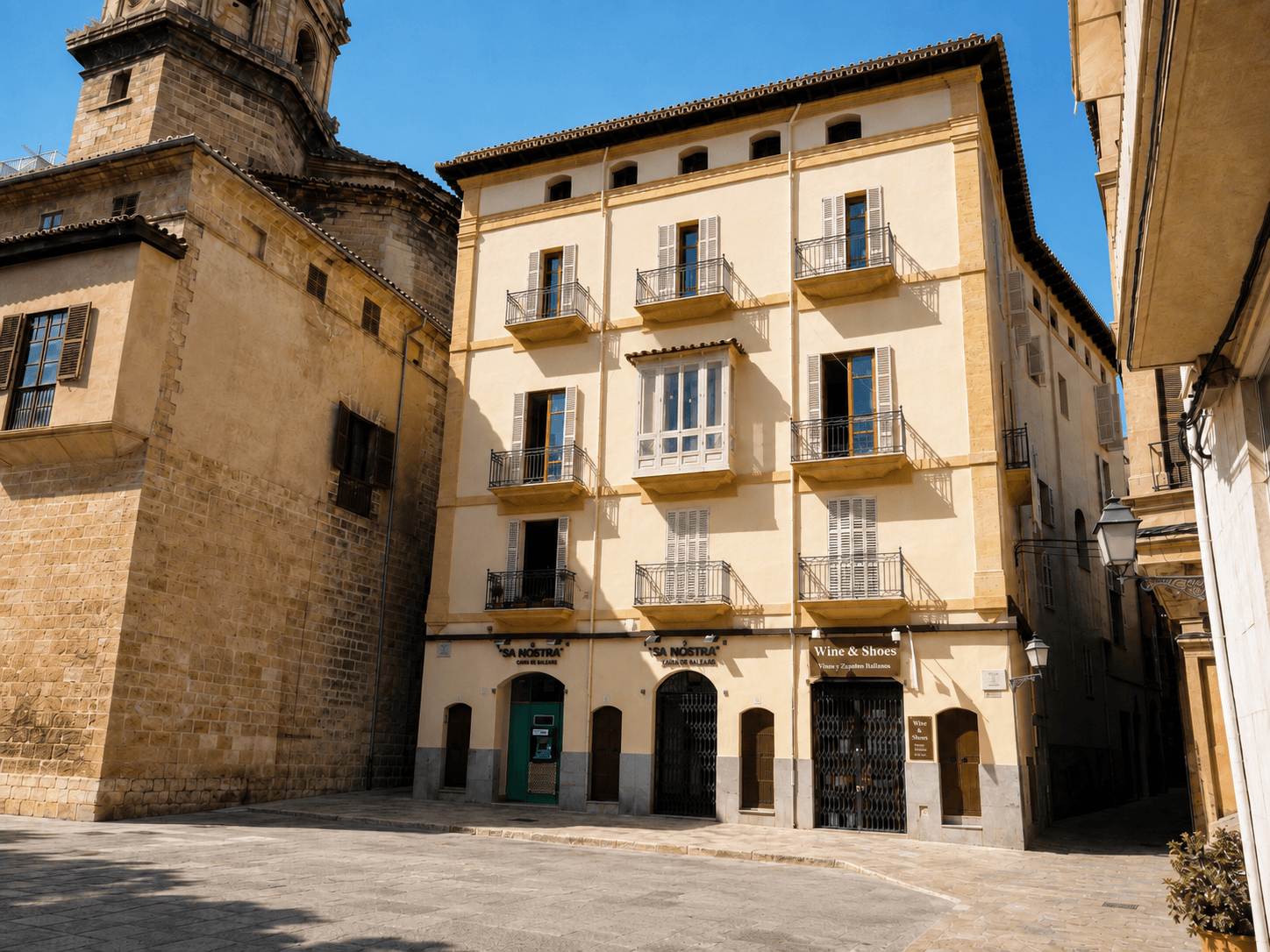 Edificio de esquina de fachada amarilla con balcones de barandilla de hierro, junto a la torre de piedra de una iglesia, en una plaza empedrada.