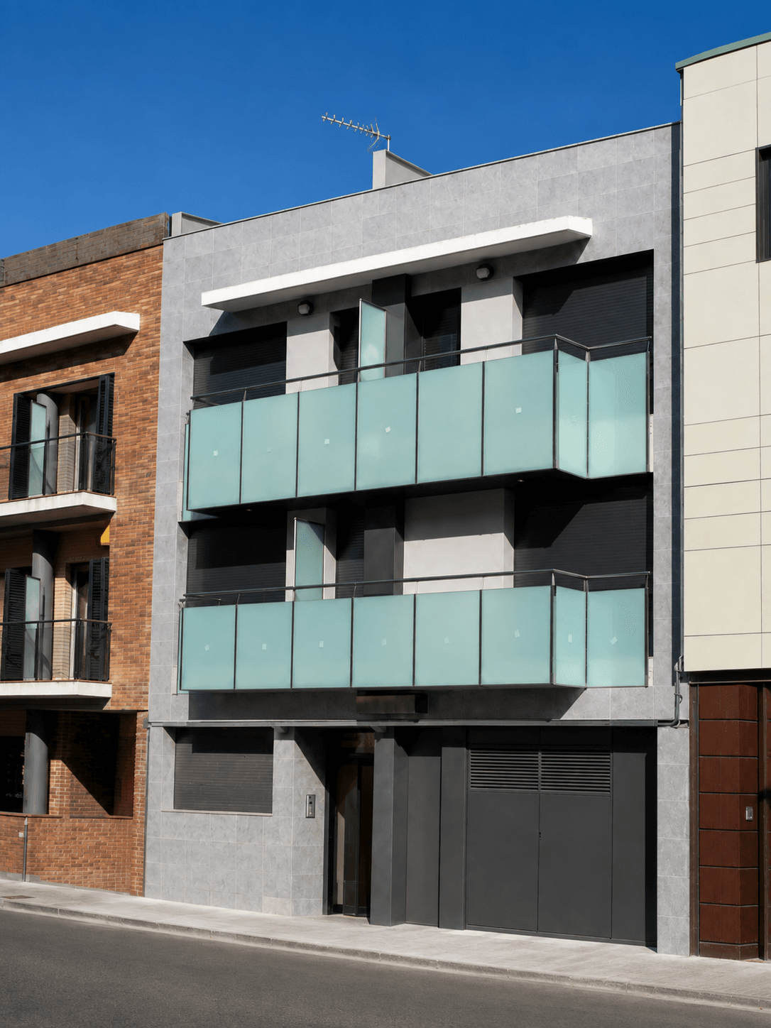 Edificio de tres plantas de fachada gris con balcones de vidrio traslúcido verdoso y puerta de garaje, entre una fachada de ladrillo y otra clara.