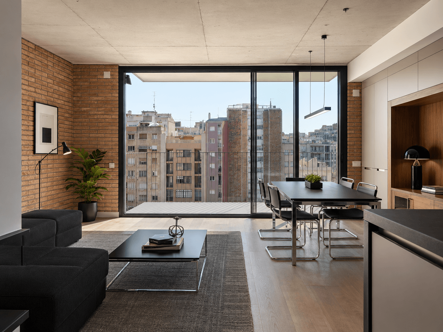 Salón comedor de un ático con ventanal a la terraza y vistas a los tejados de Barcelona, sofá oscuro, mesa negra y pared de ladrillo visto.
