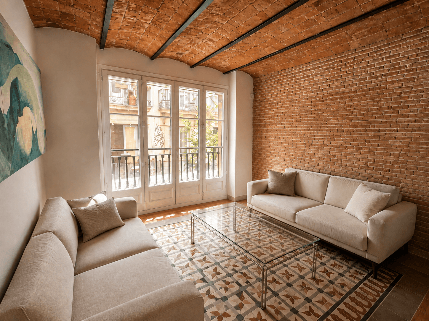 Salón con bóveda catalana de ladrillo, pared de obra vista, dos sofás claros y suelo de mosaico hidráulico frente a un balcón.