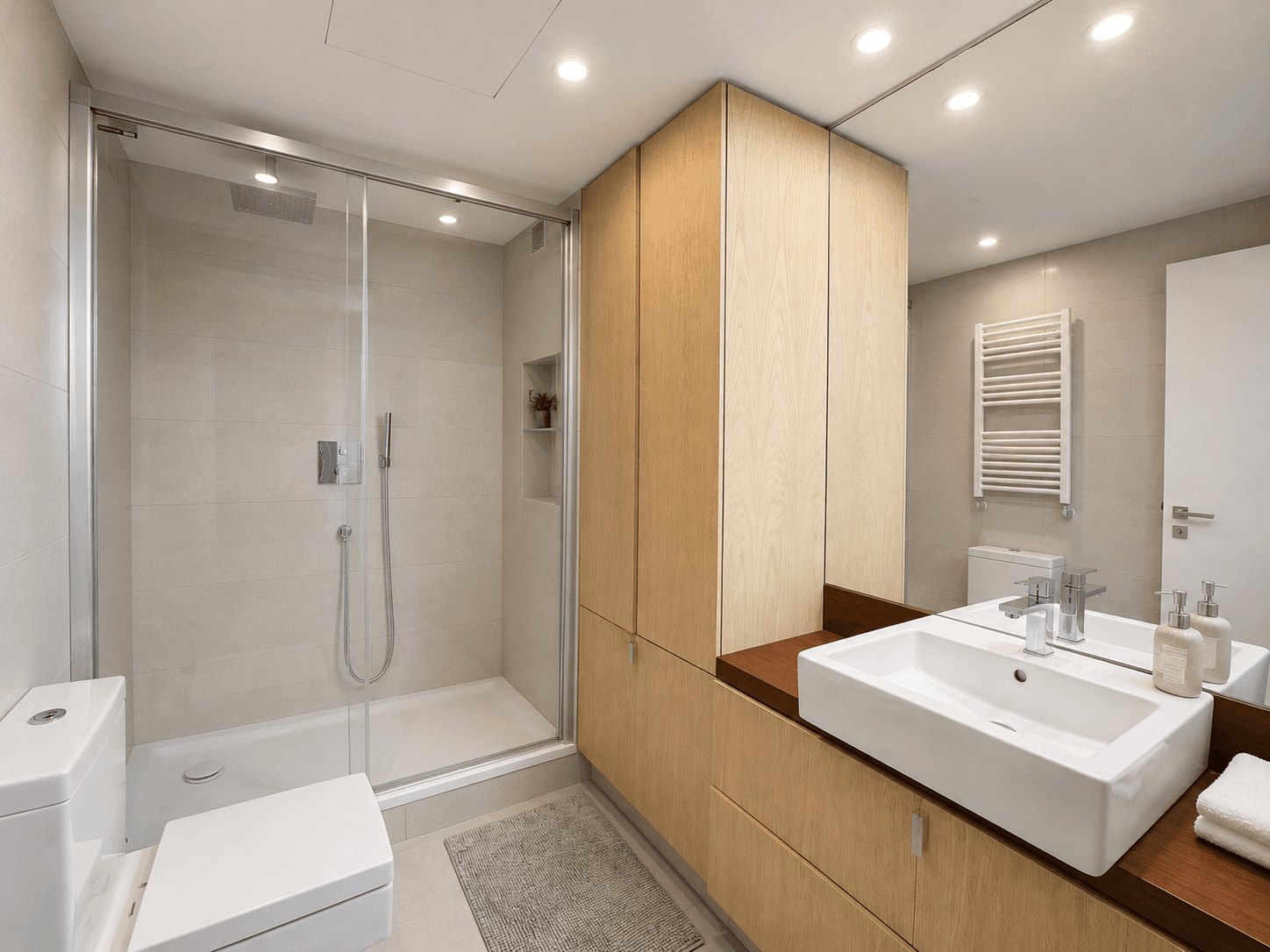 Baño con ducha de obra con mampara de vidrio, mueble de roble a medida, lavabo sobre encimera de madera y espejo de pared a pared.