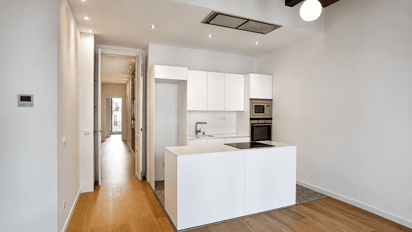 Cocina blanca con isla de encimera compacta y campana integrada en el techo, abierta al pasillo del piso.
