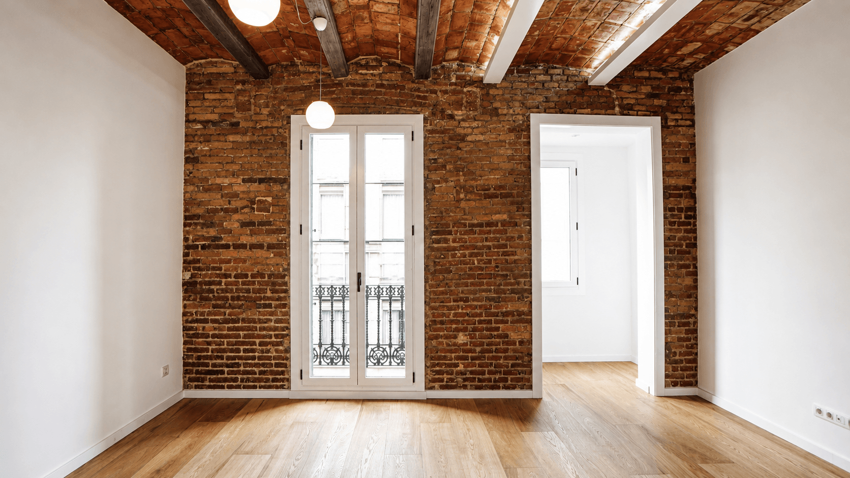 Habitación vacía con pared de ladrillo visto, bóveda catalana con vigas de madera y balcón de forja.