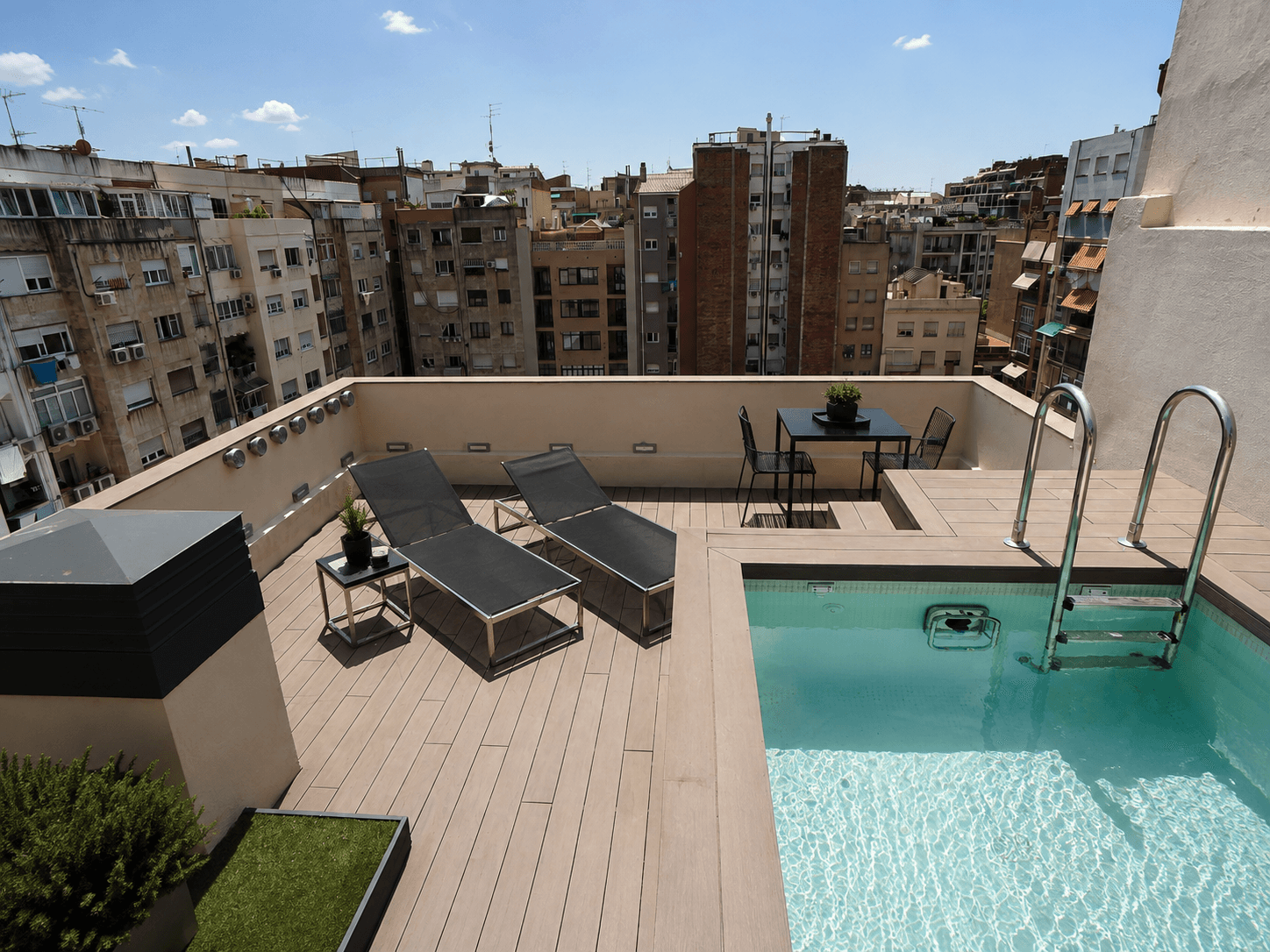 Terraza del ático con piscina, tarima, dos tumbonas y mesa alta, sobre los tejados del Eixample.