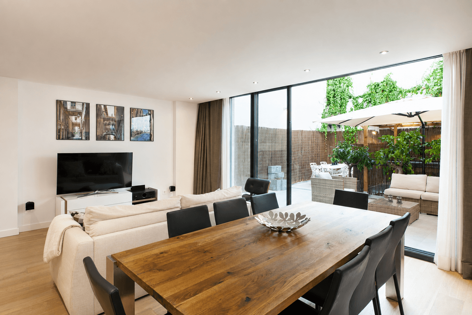 Salón comedor con mesa de roble macizo, sofá claro y ventanal corredero abierto a un patio con pérgola y plantas.