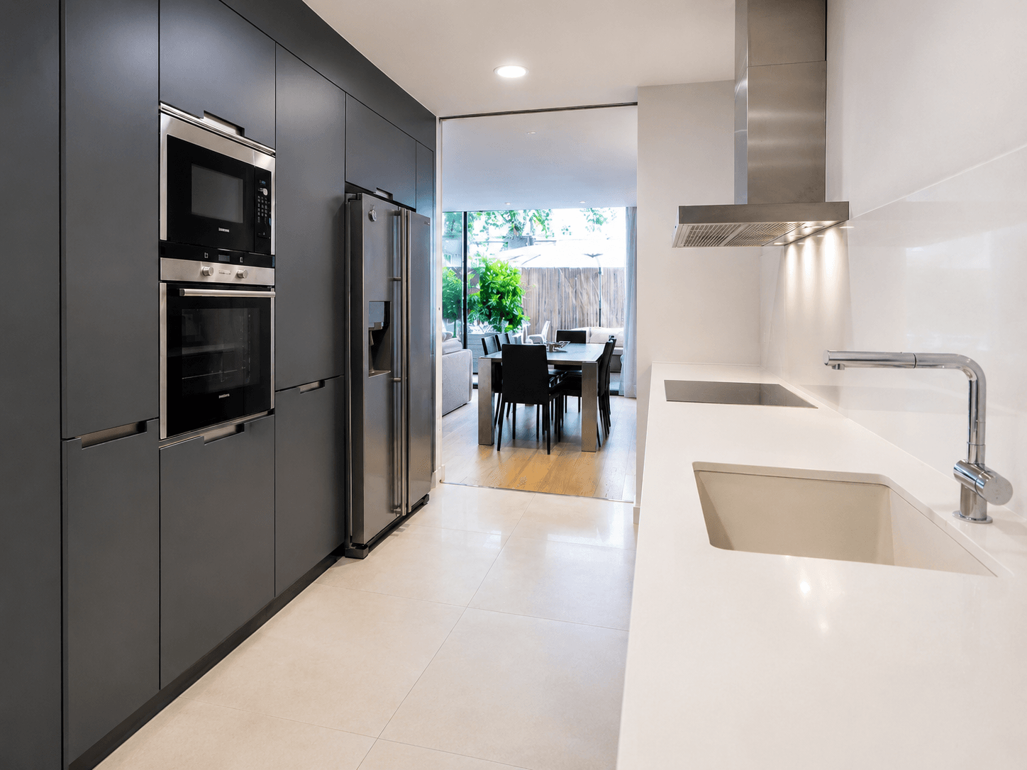 Cocina en línea con frentes gris grafito, encimera blanca continua y campana de acero, abierta al comedor y al patio del fondo.
