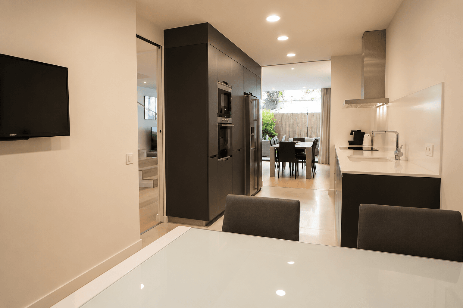 La cocina desde el office, con la mesa de comedor y el patio con cerramiento de cañizo al fondo, y la escalera del altillo tras la puerta corredera de vidrio.