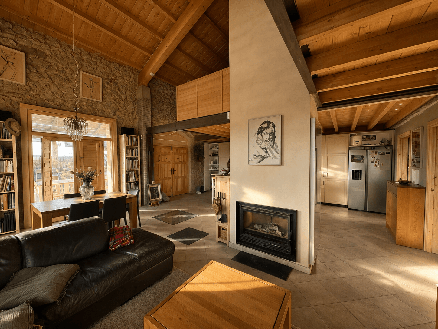 Salón comedor bajo cubierta de madera a dos aguas, con muro de piedra, chimenea de obra y la cocina al fondo.