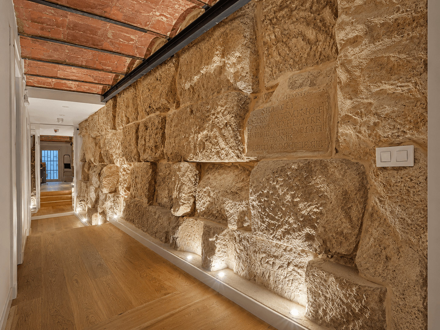 Pasillo con el muro de sillería de la muralla romana visto, iluminado desde el suelo, y techo de bovedilla con vigas de hierro.