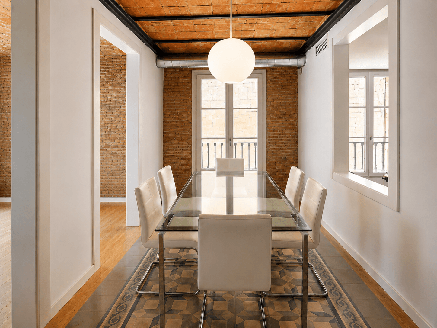 Comedor estrecho con mesa de cristal, pared de ladrillo visto, suelo de mosaico y balcón al fondo.