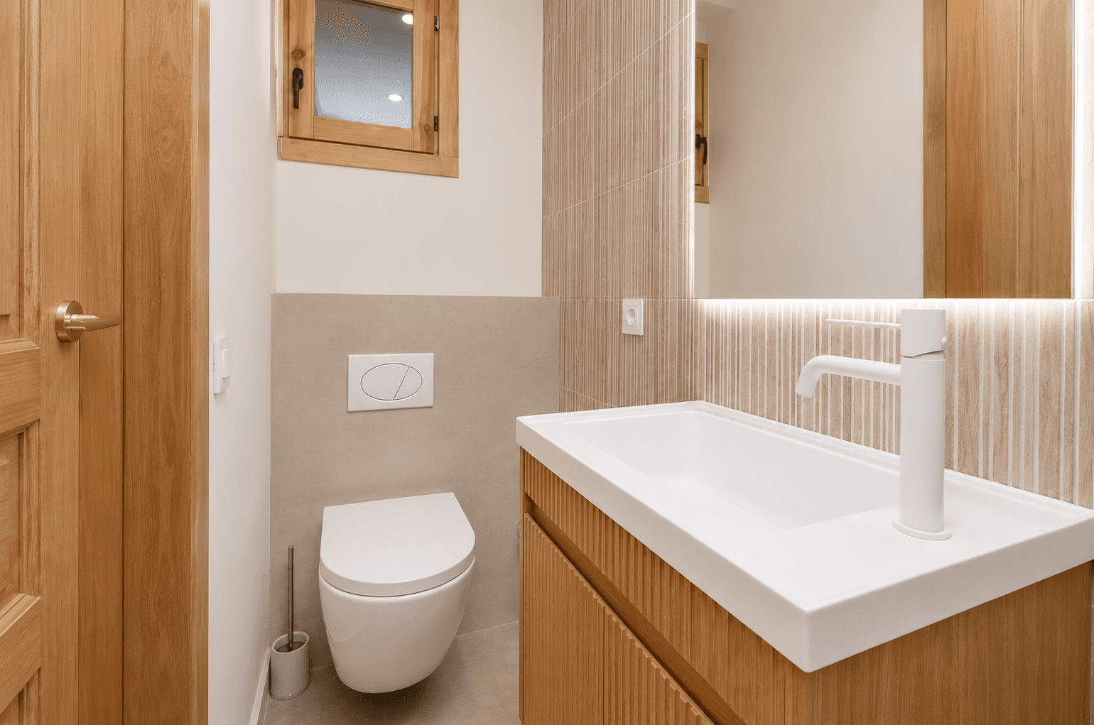 Aseo con lavabo blanco sobre mueble de roble ranurado, alicatado vertical retroiluminado e inodoro suspendido.