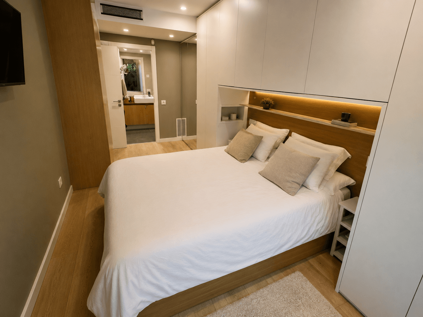 Dormitorio con cabecero de roble con luz integrada, armarios empotrados y baño en suite al fondo.
