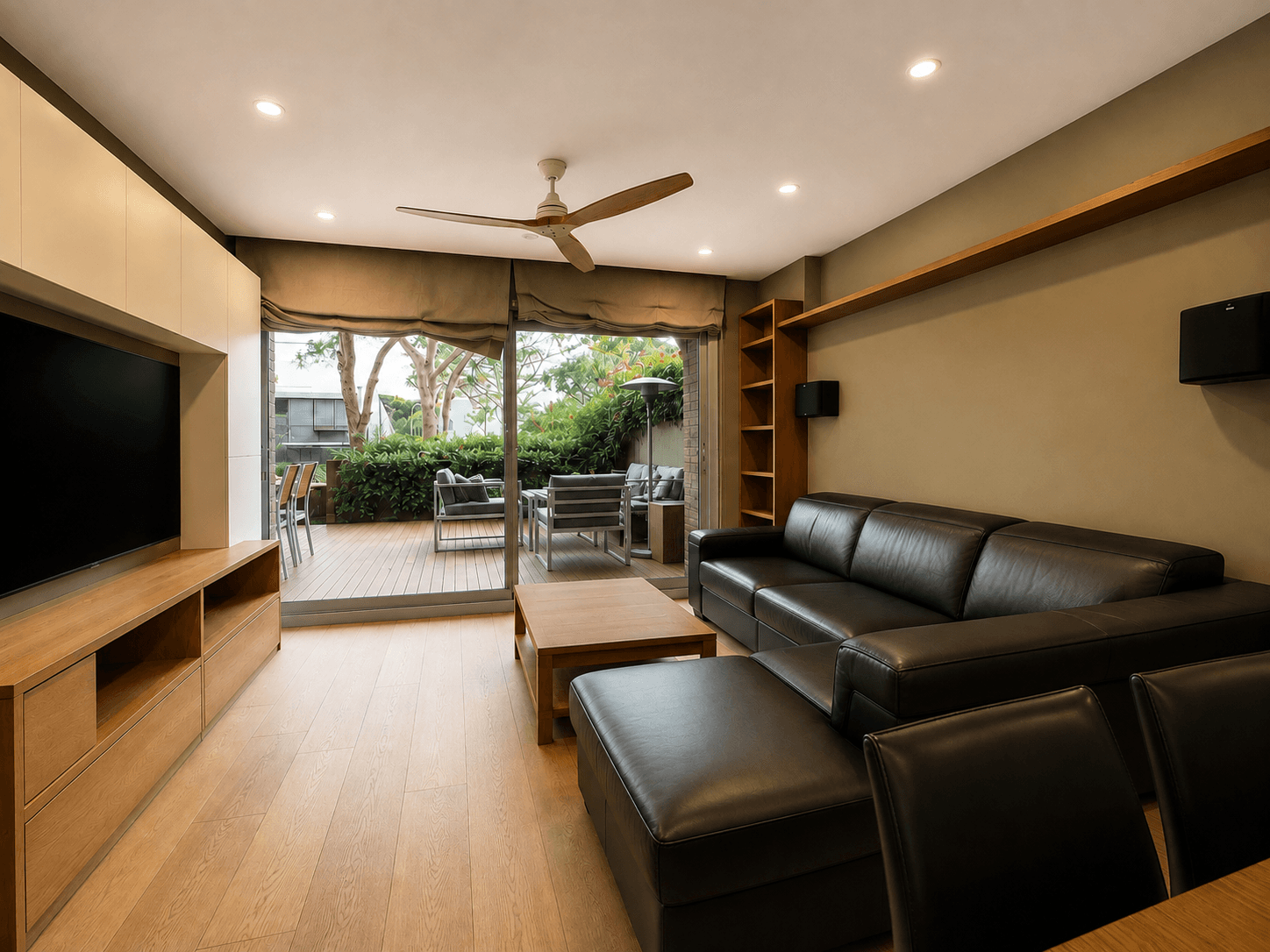 Salón alargado con mueble de televisión a medida, sofá de piel oscuro y suelo de madera clara, abierto por un ventanal corredero a una terraza de madera con jardín.