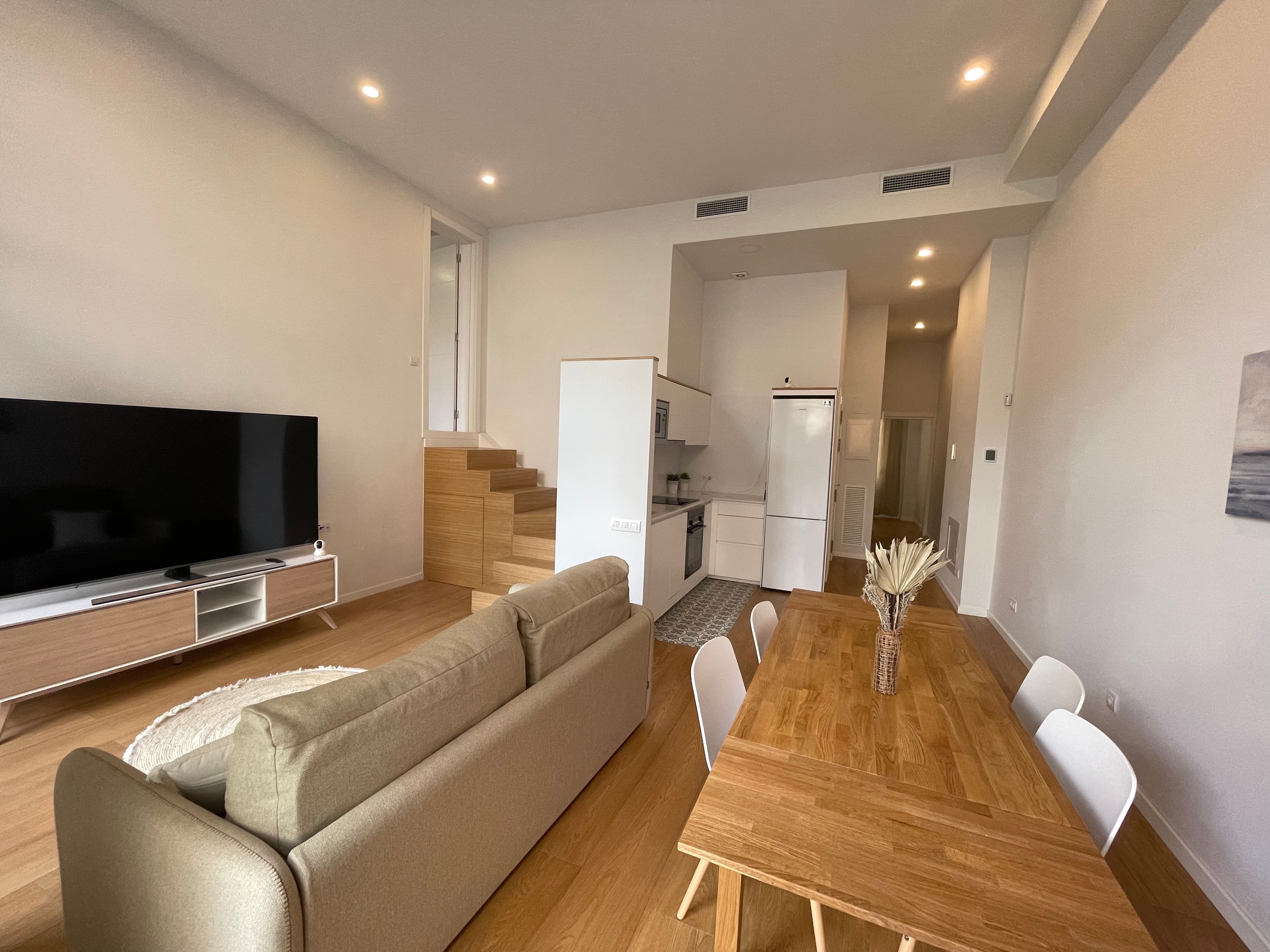 Salón alargado con cocina compacta al fondo y una escalera de peldaños de madera que sube al altillo.