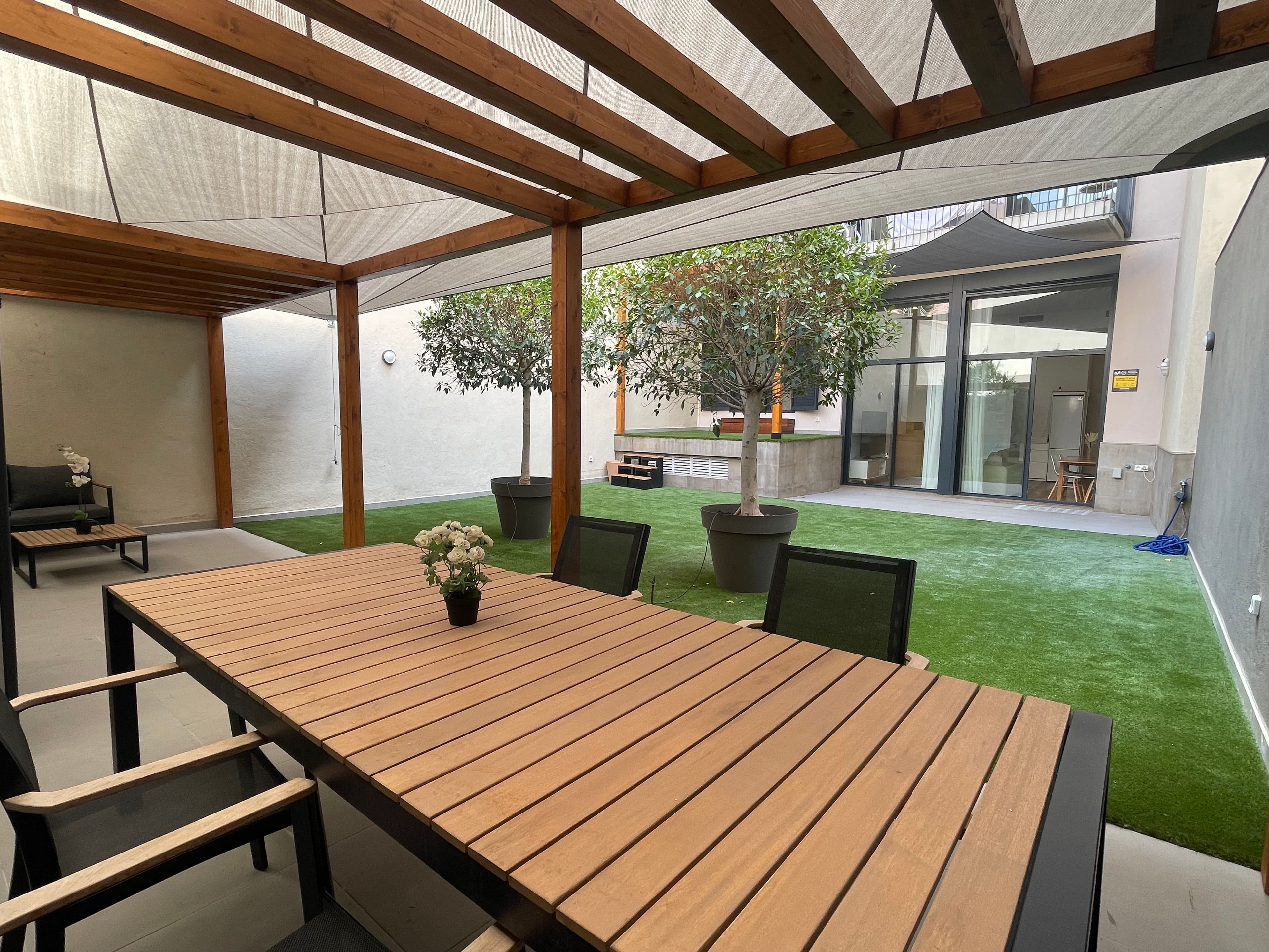 Patio con pérgola de madera y toldo, mesa larga de teca, césped y dos ficus en maceta.